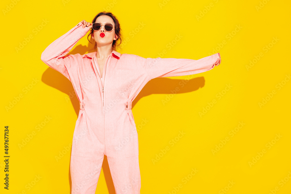Young beautiful smiling female in trendy summer pink overalls. Sexy carefree woman posing near yellow wall in studio. Positive model having fun and going crazy. Cheerful and happy. In sunglasses Young beautiful smiling female in trendy summer pink overalls. Sexy carefree woman posing near yellow wall in studio. Positive model having fun and going crazy. Cheerful and happy. In sunglasses