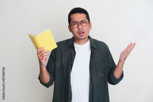 Adult Asian man looking to camera and showing confused expression while his hand holding a book photo