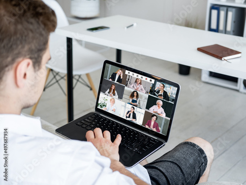 Group video call. Remote meeting. Wfh telecommuting. Relaxed man using laptop working from home online with female business team on screen.