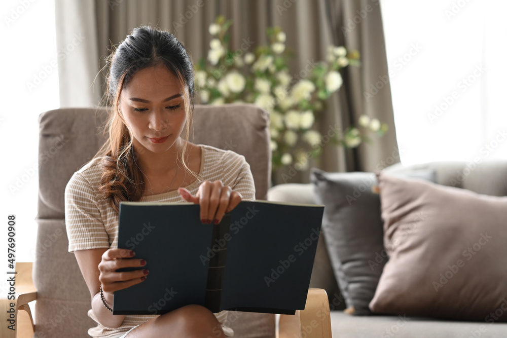 Peaceful asian woman reading book while sitting on beige armchair in her apartment. Peaceful asian woman reading book while sitting on beige armchair in her apartment.