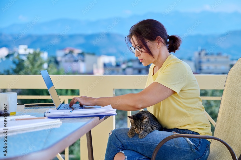 Middle-aged woman working in home office on terrace with pet cat in her arms
