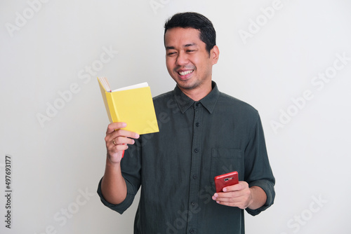Adult Asian man smiling relax while reading a book and holding mobile phone