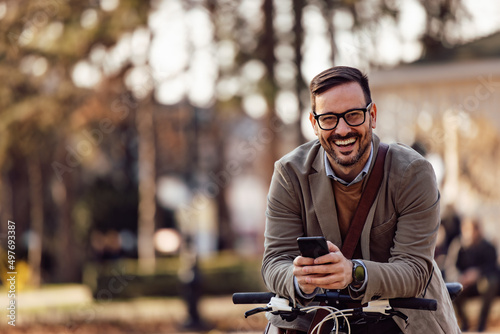 Portrait of happy caucasian man with glasses, leaning on his bike, holding his phone.