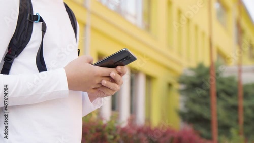 A Caucasian teenage boy looks at a smartphone - closeup on the phone - a school in the background