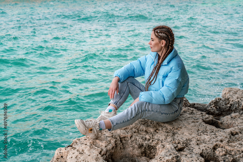 A woman in a blue jacket sits on a rock above a cliff above the sea, looking at the stormy ocean. Girl traveler rests, thinks, dreams, enjoys nature. Peace and calm landscape, windy weather.