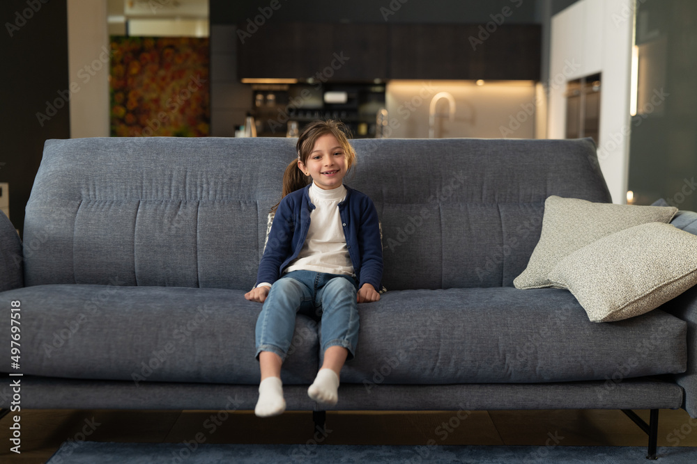 Cinematic authentic close up shot of happy cute little girl is smiling in camera while sitting on sofa in living room at home. Cinematic authentic close up shot of happy cute little girl is smiling in camera while sitting on sofa in living room at home.