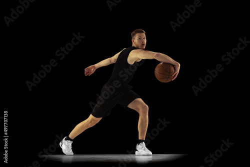 Dynamic portrait of young man, basketball player in black uniform training isolated on dark background. Achievements, sport career, motion concepts.
