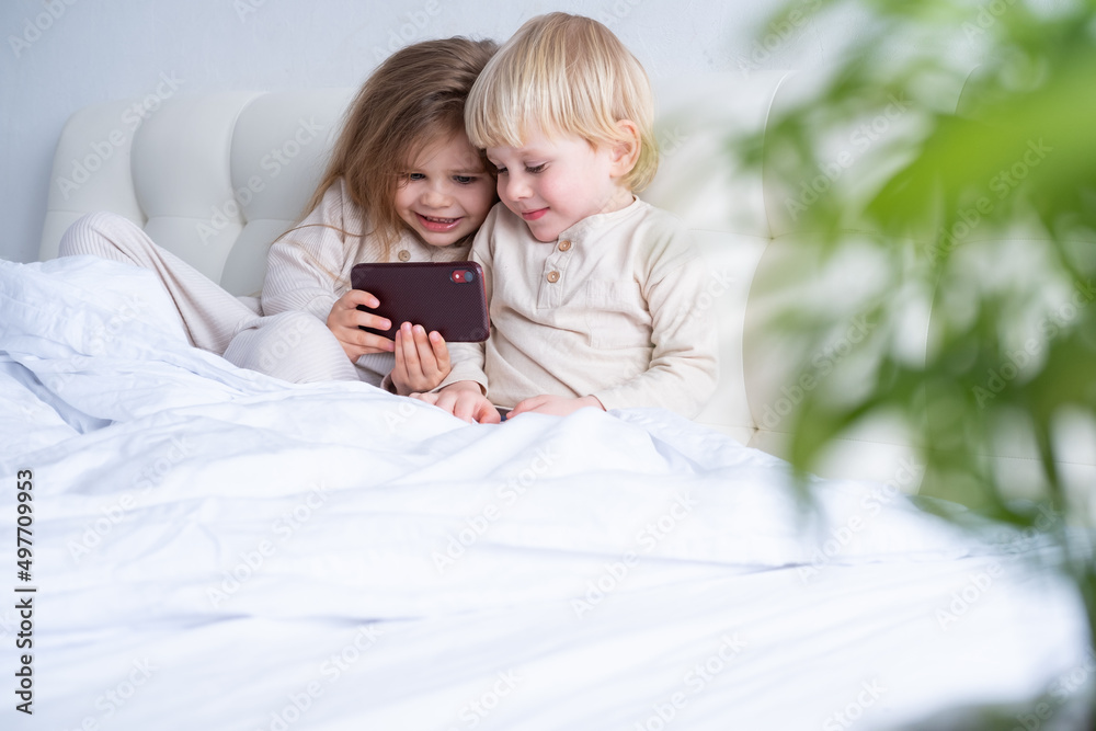 two cute kids girl and boy brother and sister using phone sitting on bed at light modern bedroom two cute kids girl and boy brother and sister using phone sitting on bed at light modern bedroom