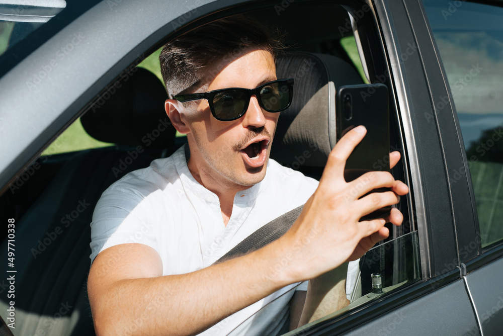 Male driver with surprised, shocked expression and an open mouth filming something on mobile phone camera while sitting in car. Caucasian man driving car and using smartphone, close-up