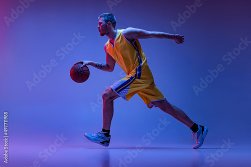 Studio shot of muscled man, basketball player training with ball isolated on purple background in neon light. Goals, sport, motion, activity concepts.