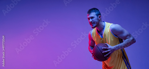 Studio shot of muscled man, basketball player training with ball isolated on purple background in neon light. Goals, sport, motion, activity concepts.