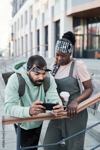 Young multi-ethnic friends leaning on railing in city and watching video on smartphone