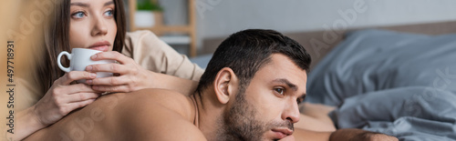 Muscular man looking away while lying near girlfriend with cup on bed, banner.