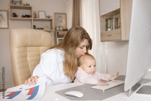 Working young mother in maternity leave work remotely or study at home on computer while infant daughter hold papers and looking at screen. Modern day mother of little child sit at desk use gadgets