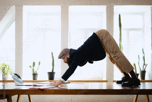 Plasticity. Young bearded man, office clerk having fun, doing yoga on wooden table in modern office. Concept of business, healthy lifestyle, sport, hobby