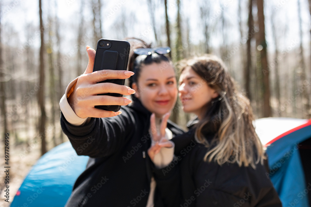 Two young woman taking selfie with smart phone in forest. One of them is making peace sign wth her hands. The focus is on girls hand an her telephone. Camping tents and hammock background. Two young woman taking selfie with smart phone in forest. One of them is making peace sign wth her hands. The focus is on girls hand an her telephone. Camping tents and hammock background.