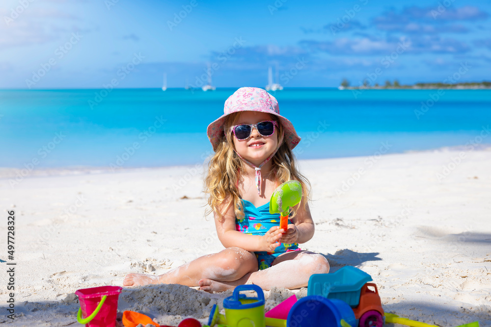 Portrait of a beautiful, blonde toddler girl sitting on a tropical beach and playing with colorful toys during her vacation time