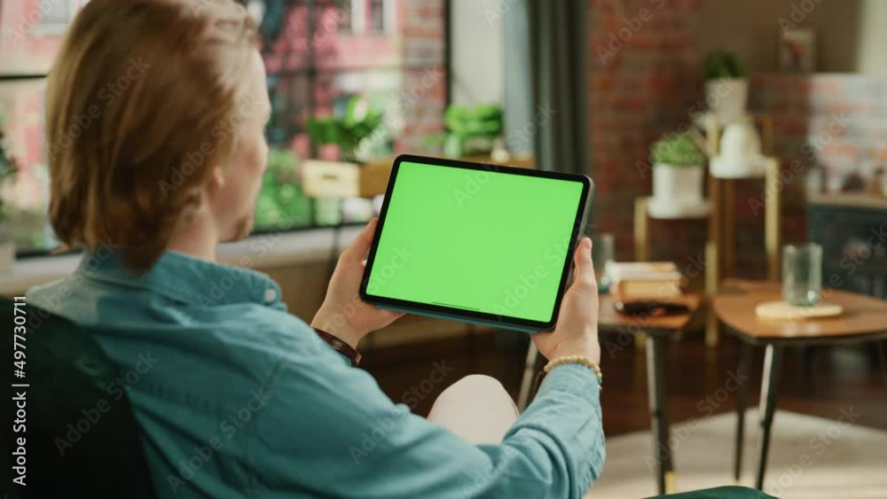 Young Man Holding Tablet Computer with Green Screen Mock Up Display. Male Relaxing at Home, Watching Videos and Reading Social Media Posts on Mobile Device. Close Up Over the Shoulder Footage. Young Man Holding Tablet Computer with Green Screen Mock Up Display. Male Relaxing at Home, Watching Videos and Reading Social Media Posts on Mobile Device. Close Up Over the Shoulder Footage.