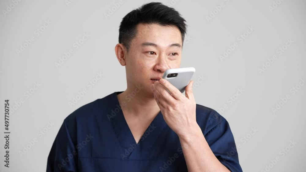 Smiling Asian doctor recording voice message by phone in the grey studio Smiling Asian doctor recording voice message by phone in the grey studio
