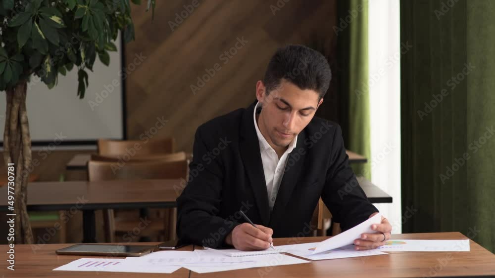 Concentrated young arab businessman looking through marketing research report, analyzing statistical data in charts, developing growth strategy, working alone Concentrated young arab businessman looking through marketing research report, analyzing statistical data in charts, developing growth strategy, working alone