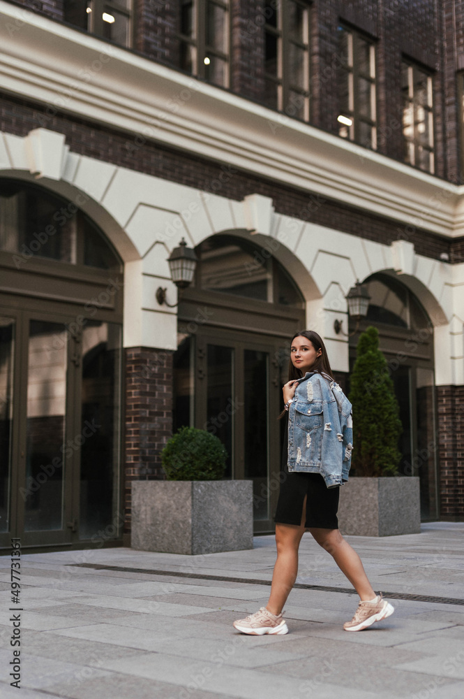 A girl in a black dress-sneakers in a denim jacket on a city street on a sunny clear day walks posing for the camera in the courtyard of an office building