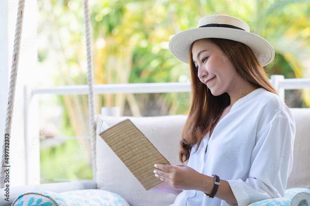 Portrait image of a beautiful young asian woman with hat reading book while sitting and relaxing on swing sofa