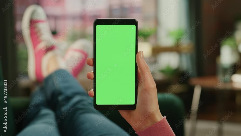 Feminine Hand Holding a Smartphone with Green Screen Mock Up Display. Female is Relaxing on a Couch at Home, Watching Videos and Reading Social Media Posts on Mobile Device. Close Up POV Footage.