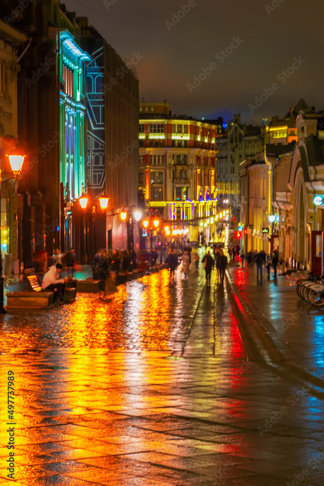 Abstract bright blurred background with people city street in rainy night. Vivid illumination, reflection, street lamps