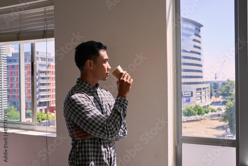 A young businessman drinks his coffee and looks out on the cityscape from office photo