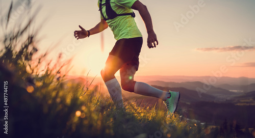 Cropped photo of Middle-aged mountain trail runner man dressed bright t-shirt with a backpack endurance running uphill by picturesque hills at sunset time. Sporty active people concept image. photo