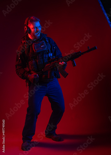 a fighter in equipment, body armor, headphones holds a machine gun, aims at the target, hits the target through the sight. military business concept. red background. blue-red light