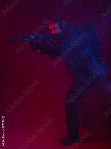 soldier in full gear with weapons. a man in headphones  body armor  with a backpack and a belt. red background. colored  blue-red light. smoke around the military. explosion  chemical attack