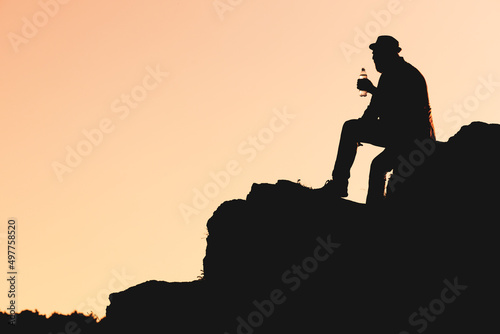 silhouette of a man in a hat who drinks water from a bottle while sitting on a mountainside