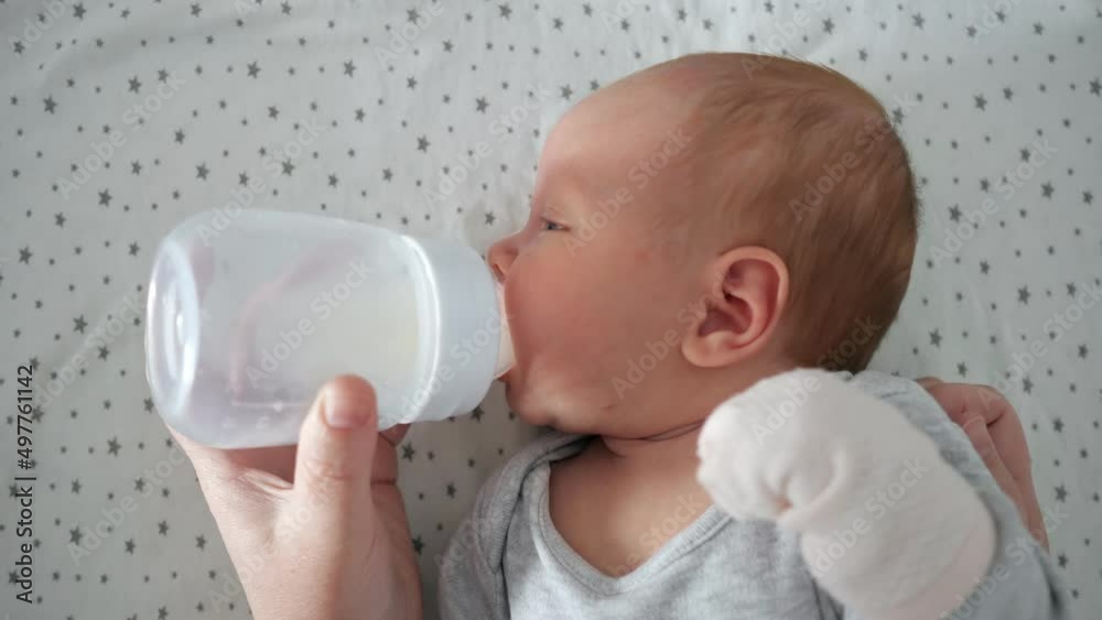 Newborn baby girl with half closed eyes drinks water from plastic bottle lying on bed. Mother feeds cute little kid at home close upper view Newborn baby girl with half closed eyes drinks water from plastic bottle lying on bed. Mother feeds cute little kid at home close upper view