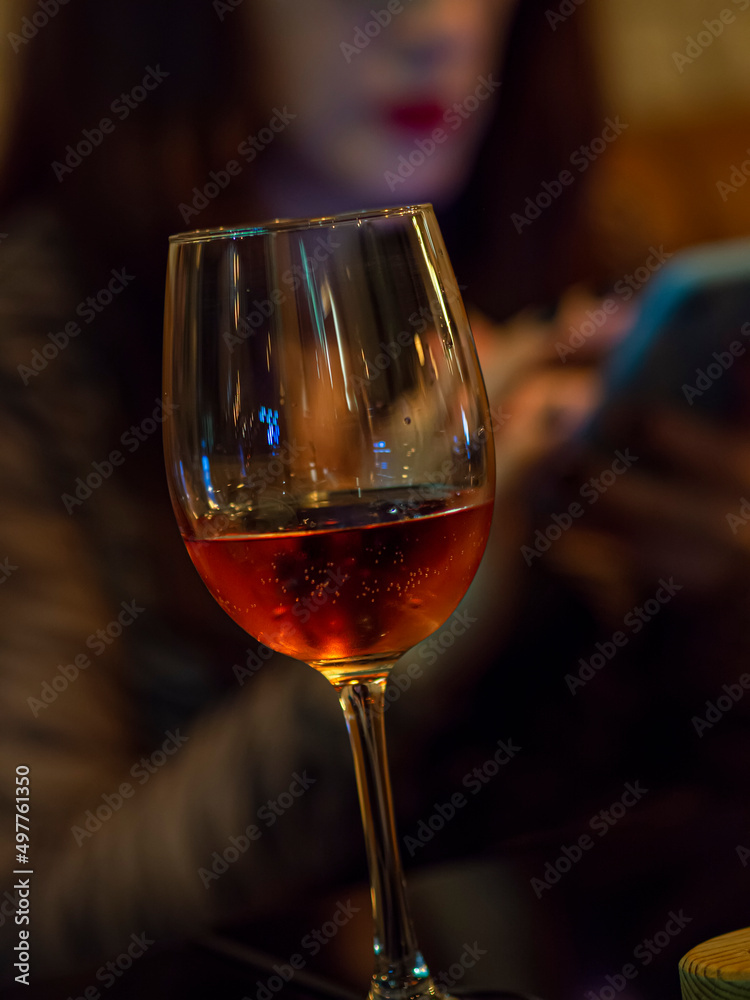 Rose wine glass with woman blurred in the background with cellphone. Woman waiting in a bar for a date while drinking. Brunette with phone in a restaurant alone.