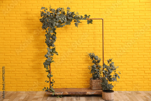 Stylish photo zone with wooden frame and green leaves near yellow brick wall indoors