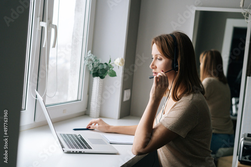 Side view of focused female operator talking using headset and consulting client sitting at window sill with laptop from home office. Freelance working having call video conference with customer.