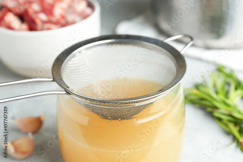Glass jar with delicious bone broth and sieve on table, closeup photo