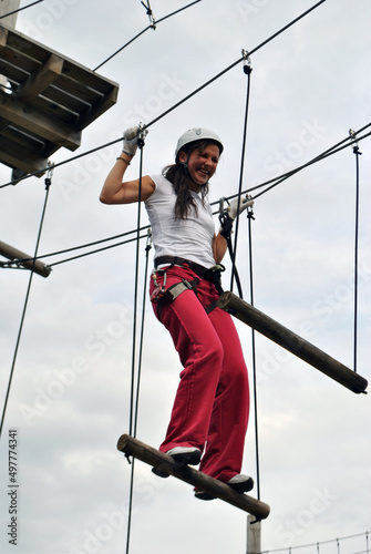 A young woman in an amusement park walks along a rope bridge in a safety harness and a sports helmet