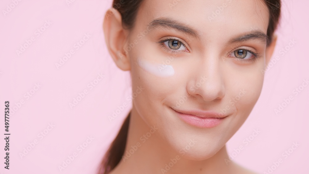 Obraz premium Close-up portrait of young attractive beauty model with perfect skin with cream smear on her face looks at the camera and smiles on light pink background | Skin care product advertising concept.
