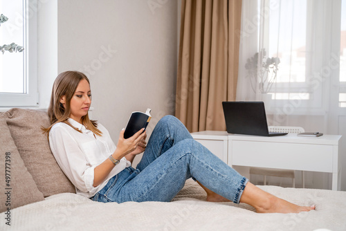 White cozy bed and a beautiful girl in a white shirt and jeans reading a book, the concept of home and comfort. Against the background of the living room, computer and window.