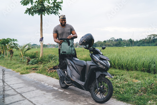 Cheerful African American tourist smiling near modern scooter while searching something in backpack at Bali, happy dark skinned hipster guy with black moped enjoying summer vacations in Indonesia