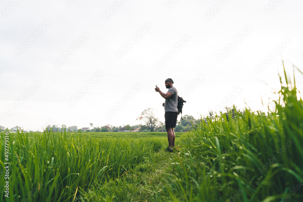 Side view of hipster guy with backpack using mobile device for clicking scenery view during summer trip to Bali, African American male blogger with smartphone shooting video content at rice fields