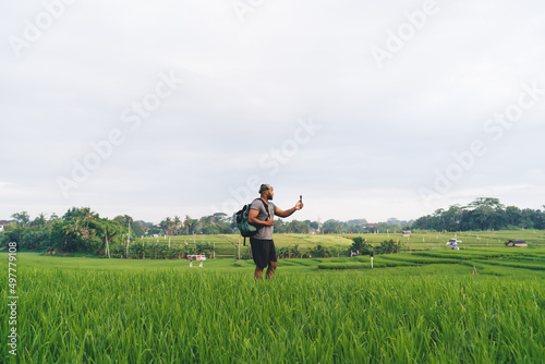 Young tourist with backpack visiting rice fields in Indonesia using modern gopro camera for shooting vlog content during summer getaway vacations, millennial hipster guy with waterproof device