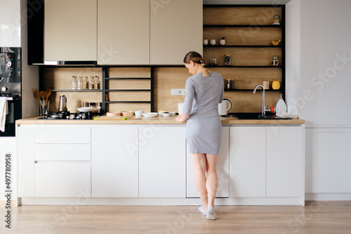 Young woman with beautiful body standing next to a modern kitchen while cooking. Back view