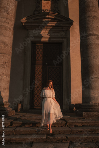 beautiful young girl with red curly hair in a long white dress barefoot walks up the old stairs near the old gray building with columns of ancient architecture at sunset, foreground
