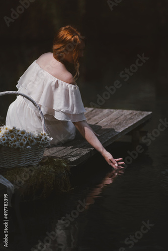 beautiful young girl with red hair in a white dress sits on a wooden pier and touches his hand to the water near her white straw basket with daisies, sunny day side view photo