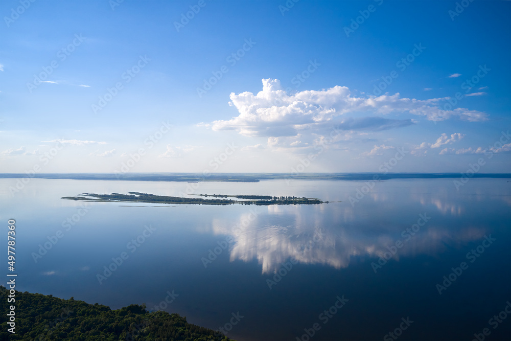 A full-flowing flat river with forested banks. There is a small island in the middle of the river. The white cloud is beautifully reflected in the water. Shooting from a drone.