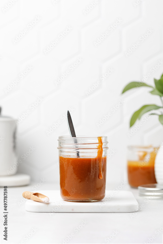 Homemade caramel in a jar with spoon. Salted Caramel sauce on white board, tile background Homemade caramel in a jar with spoon. Salted Caramel sauce on white board, tile background