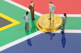 On the surface with the image of the flag of South Africa are bitcoins and miniature figures of people.